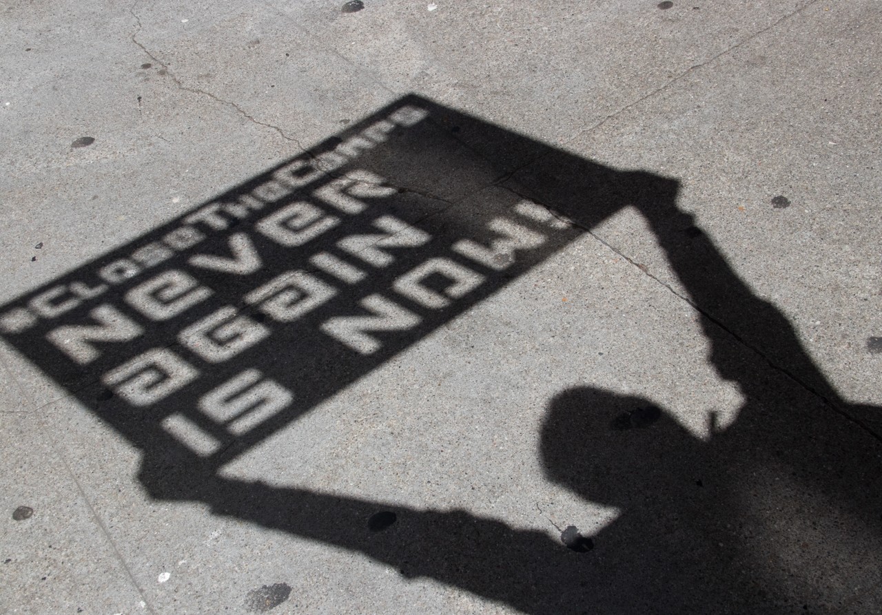 Ein Schatten, der ein Schild hält mit der Aufschrift "Close the Camps. Never again is now"
