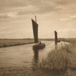 Ein sepiafarbenes Schwarz-Weiß-foto. Darauf ein Fluss, leicht gebogen dem Fotografen entgegen. Links und rechts Schilf. Auf dem Fluss zwei alte kleine Segelboote mit rechteckigem Segel.