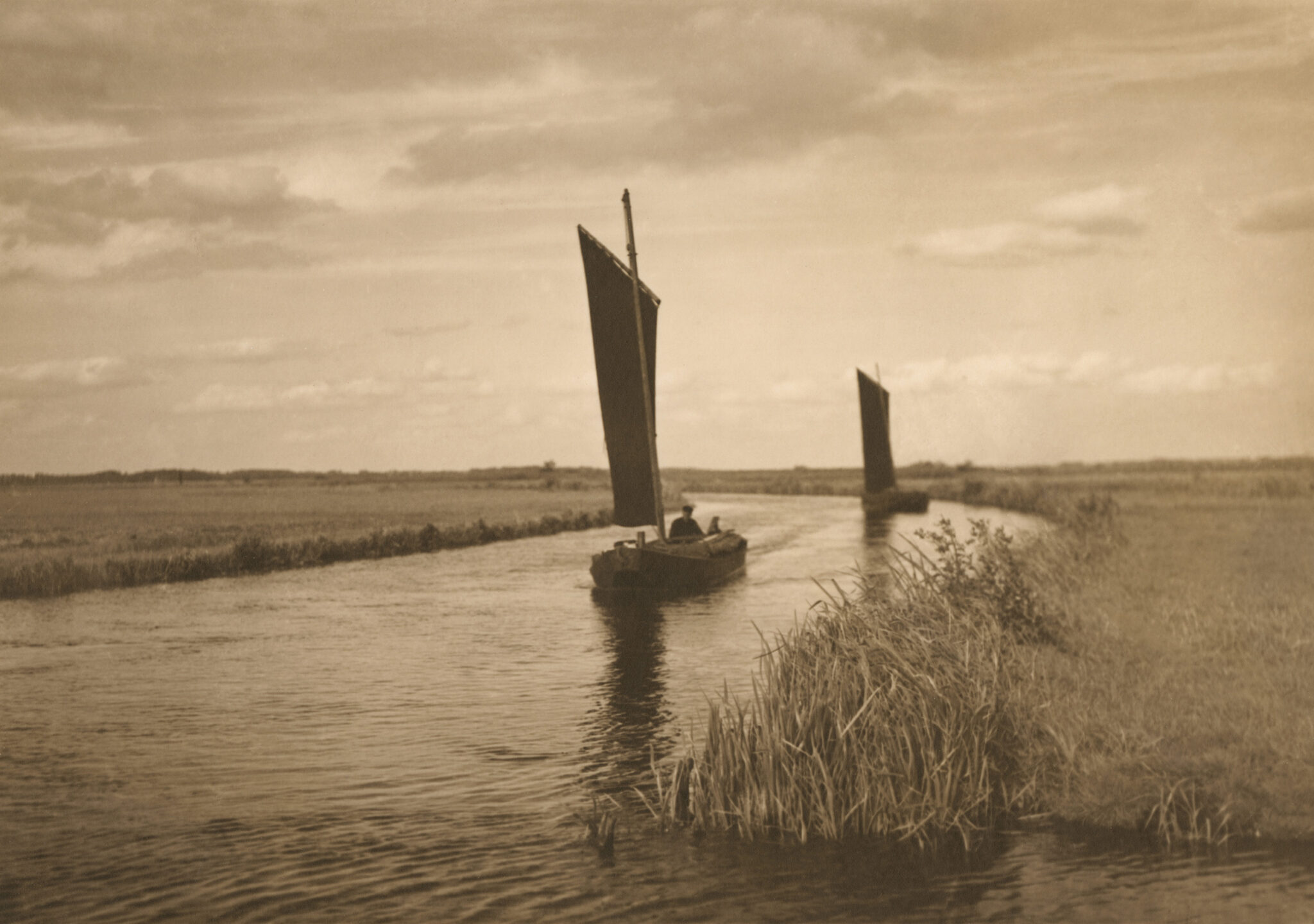 Ein sepiafarbenes Schwarz-Weiß-foto. Darauf ein Fluss, leicht gebogen dem Fotografen entgegen. Links und rechts Schilf. Auf dem Fluss zwei alte kleine Segelboote mit rechteckigem Segel.