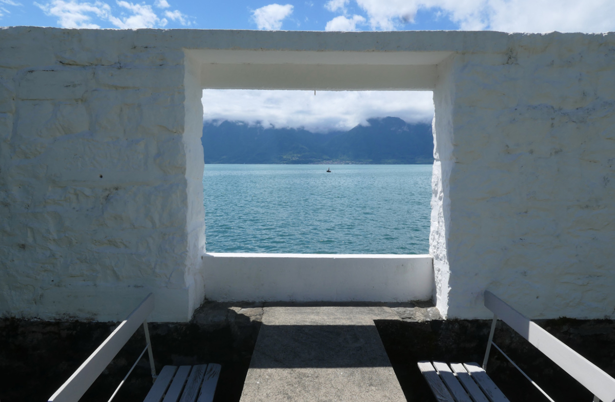 Eine weiße Mauer vor einem See. Die Mauer ist hoch, in ihr befindet sich ein offener Fensterausschnitt. Dafür zwei Bänke und ein Tisch.