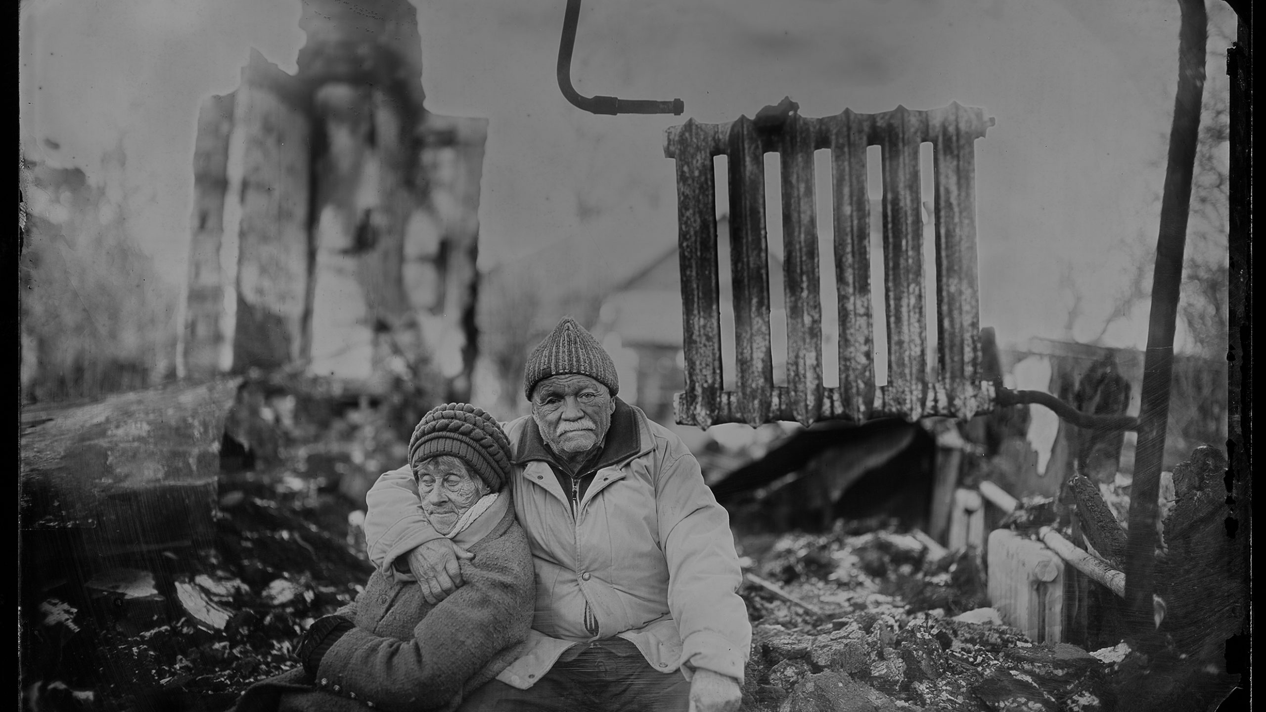 Ein Schwarz-weißes Foto, im Vordergrund scharf, ein altes Paar, ein Mann und eine Frau. Der Mann an den Arm um die Frau gelegt, sie lehnt an ihm. Im HIntergrund Ruinen. eine Heizung steht frei in der Luft, gehalten von einem Rohr.