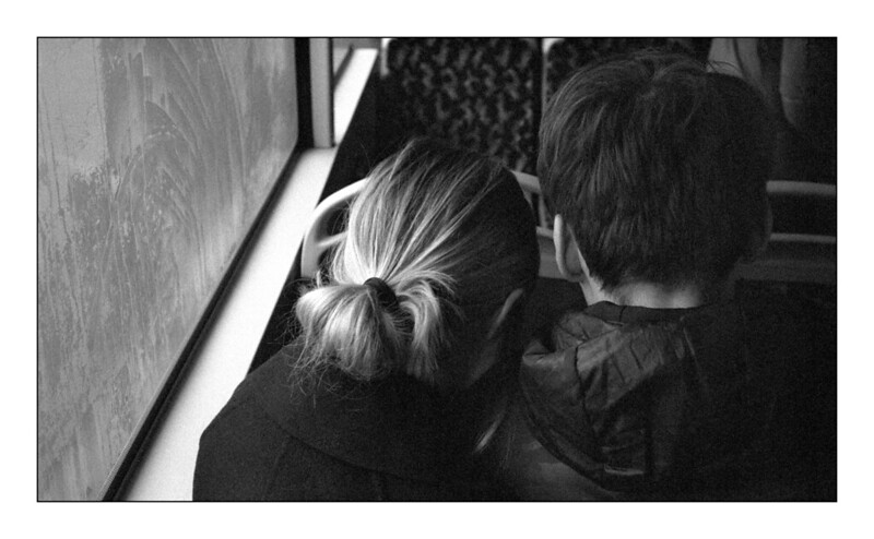 Zwei Personen von hinten, die nahe beieinander sitzen. Die Person links hat helles, zu einem Dutt gebundenes Haar, die Person rechts trägt eine dunkle Jacke. Links im Bild ist ein Fenster mit sichtbarer Fensterscheibe mit Eis darauf.